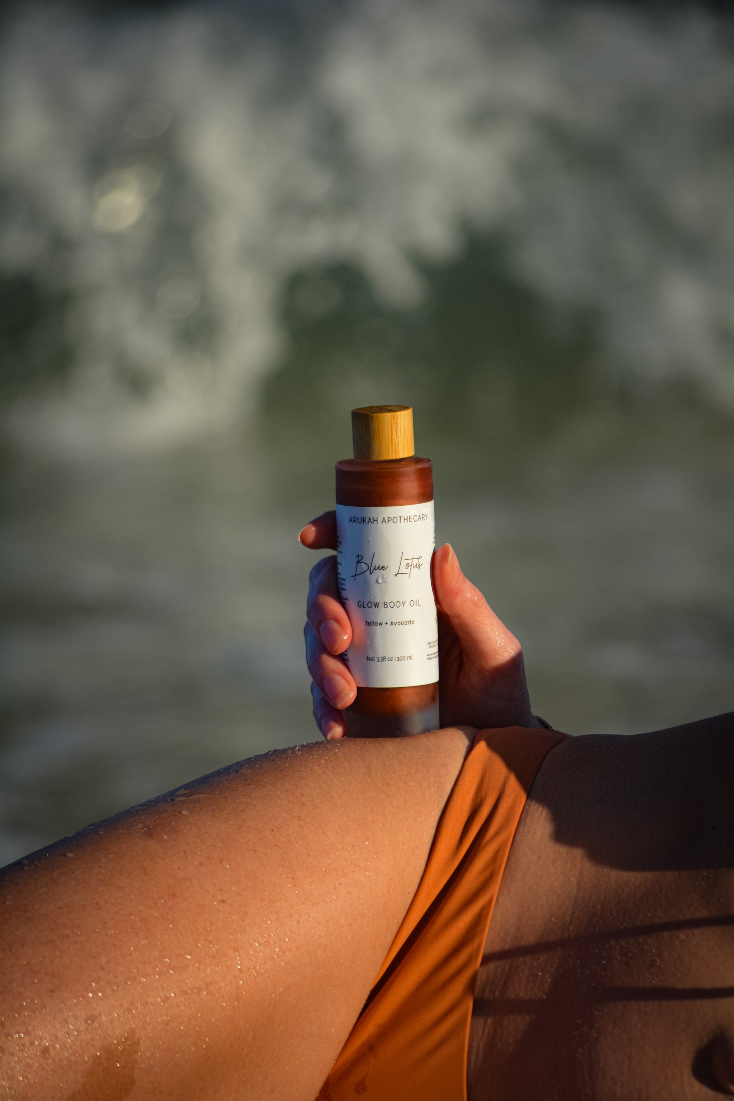 Blue Lotus Glow Body Oil held at the beach with waves in background
