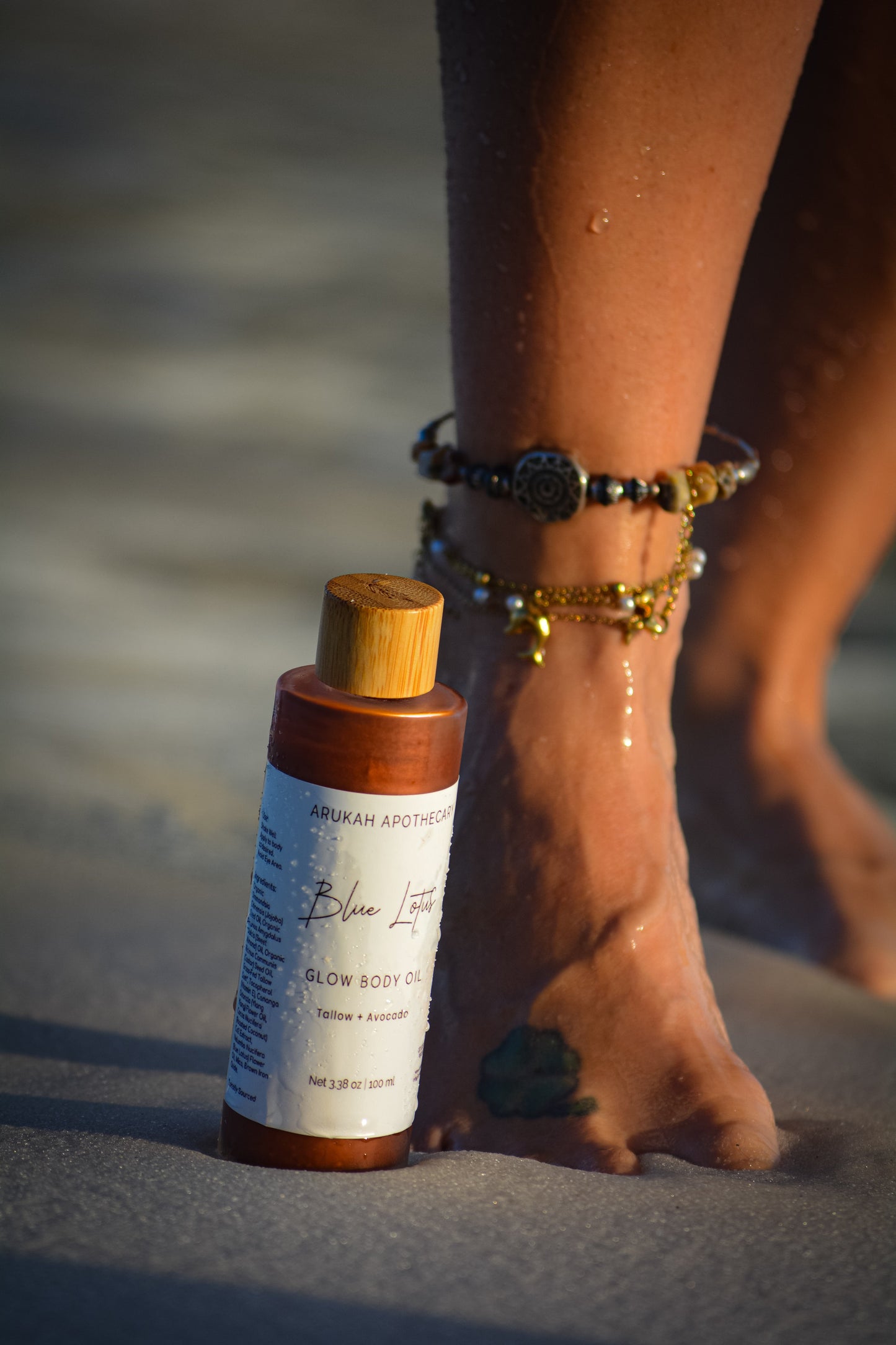 Blue Lotus Glow Body Oil on sand next to anklet-adorned feet